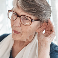 Older woman wearing glasses holding her hand to her ear as if struggling to hear.