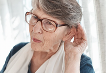 Older woman wearing glasses holding her hand to her ear as if struggling to hear.